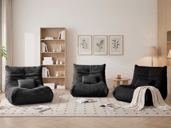 Two Zoovee's dark grey and one black ribbed corduroy lounge bean bag chairs arranged on a patterned rug in a bright, modern living room with a wooden bookshelf, wall art, and a small side table.