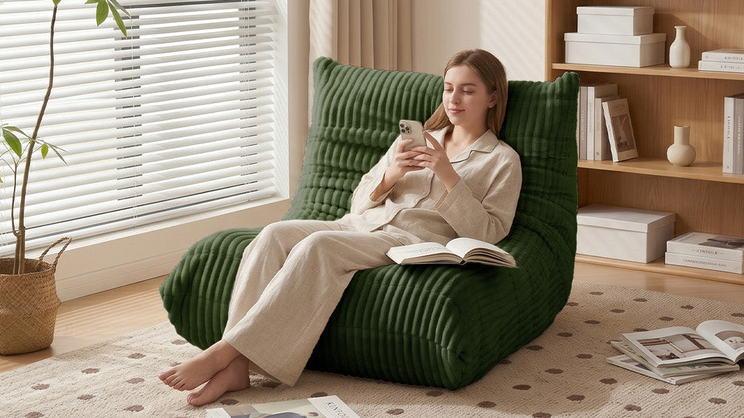 A woman in a beige pajama set relaxes in Zoovee's green ribbed lounge bean bag chair inside a bright, modern room with a wooden bookshelf, window blinds, and a potted plant.